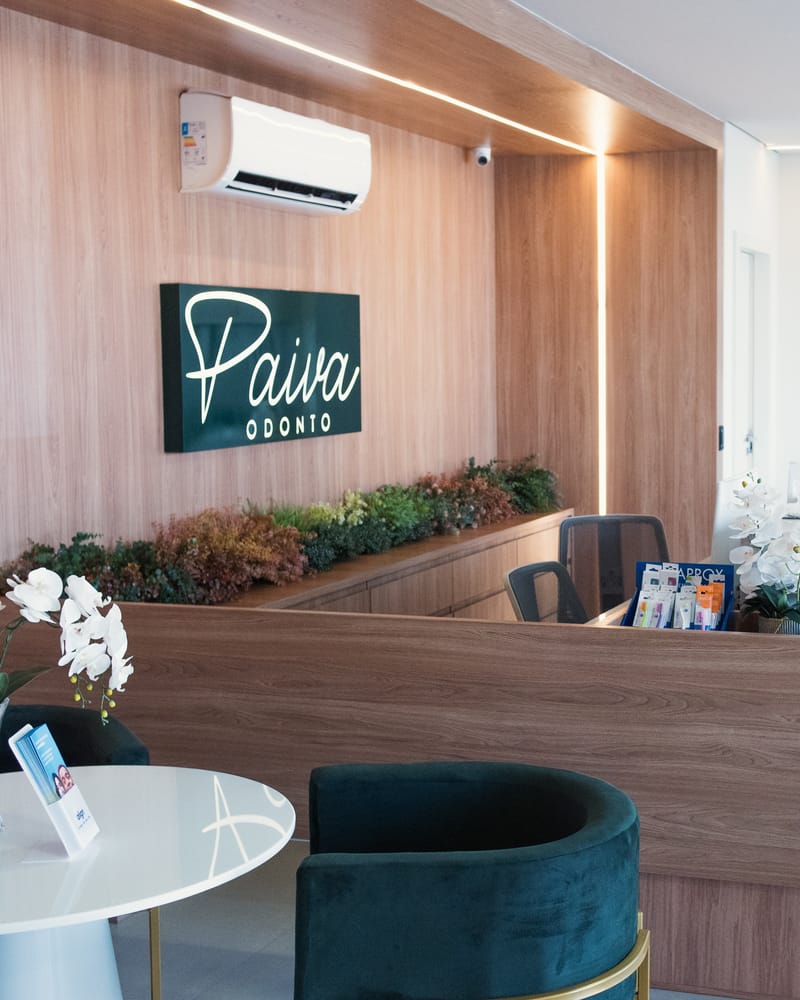Recepção da clínica Paiva Odonto em Linhares, com o letreiro da clínica no painel de madeira e poltronas de veludo verde